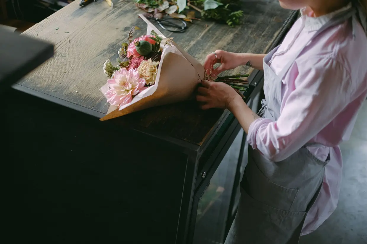 Des mains qui emballent un bouquet dans du papier kraft, sur un établi.